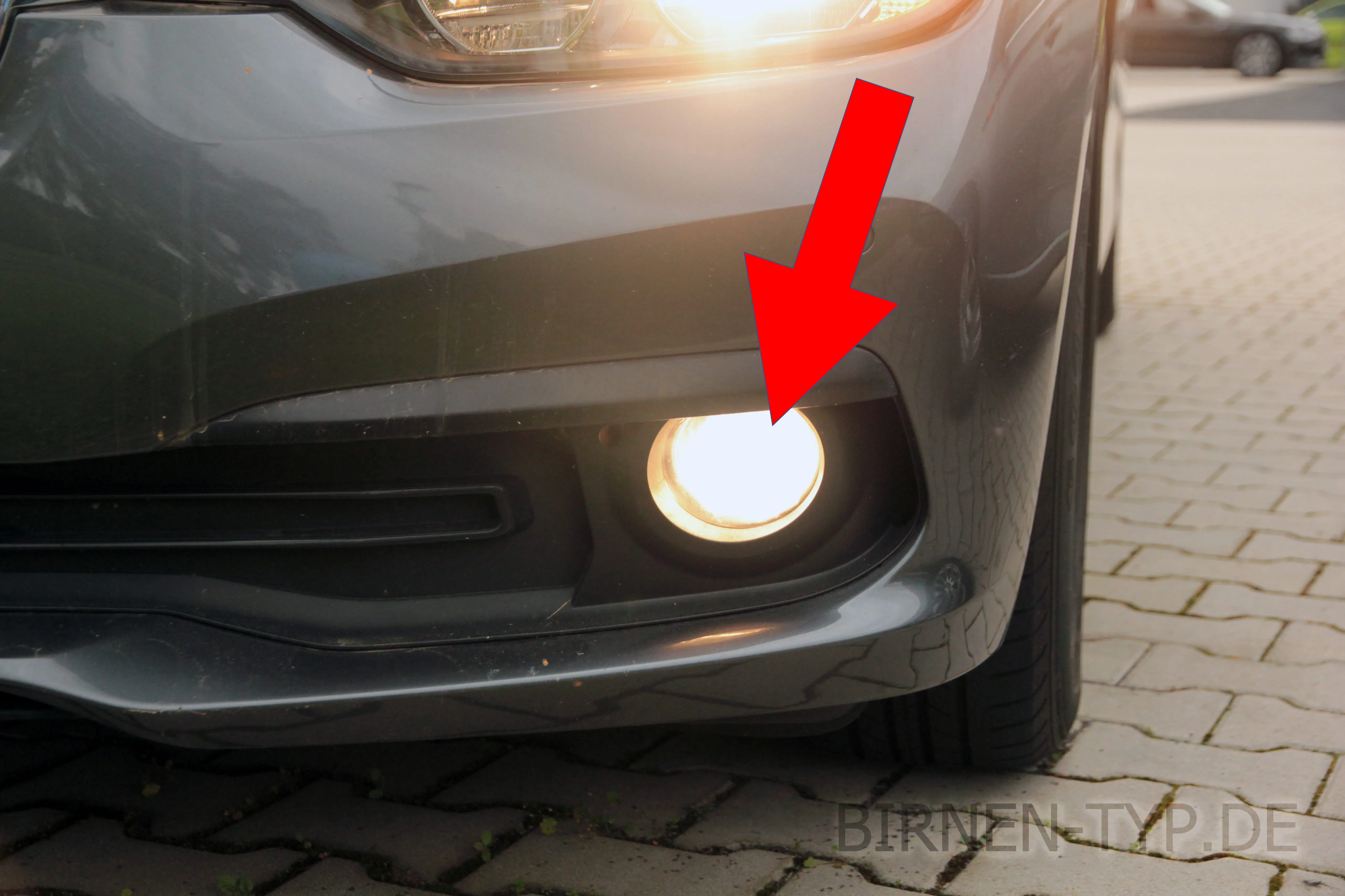 Eingeschalteter Nebelscheinwerfer mit H8-Halogenbirne in der Frontschürze eines grauen BMW 3er F30, ein roter Pfeil zeigt auf die Leuchte